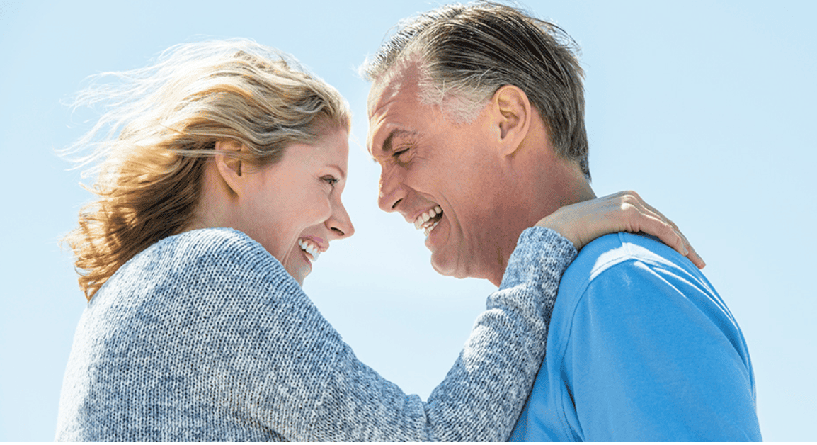 A joyful middle-aged couple embracing and laughing outdoors under a bright sky, symbolizing love, connection, and emotional well-being.