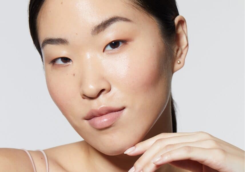 A close-up portrait of a woman with smooth, glowing skin, showcasing a natural and healthy complexion. She has her hair tied back and gently rests her hand near her chin, emphasizing her radiant, well-nourished skin.