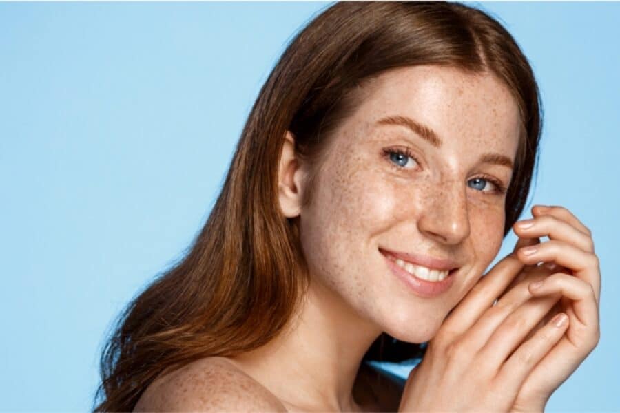 A close-up of a young woman with light freckles, smooth skin, and bright blue eyes, smiling gently while posing with her hands near her face against a soft blue background.