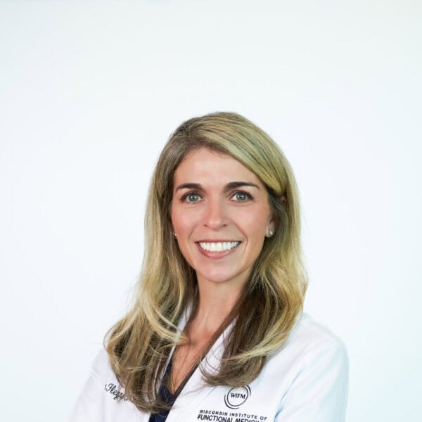 A woman with medium-length brown hair, wearing a purple top and smiling, standing in front of a white background.