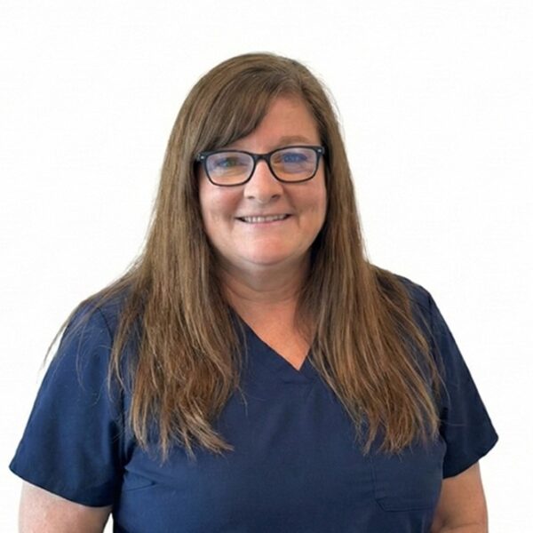 A woman with long brown hair and glasses, smiling while wearing blue scrubs against a white background.