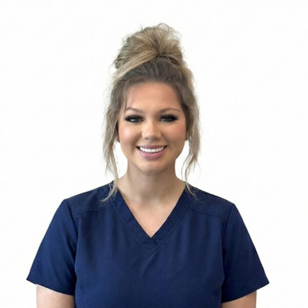 A woman with long brown hair and glasses, smiling while wearing blue scrubs against a white background.