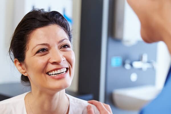 A smiling woman in a white blouse sitting in a medical setting, with a healthcare professional touching her shoulder in a supportive gesture.