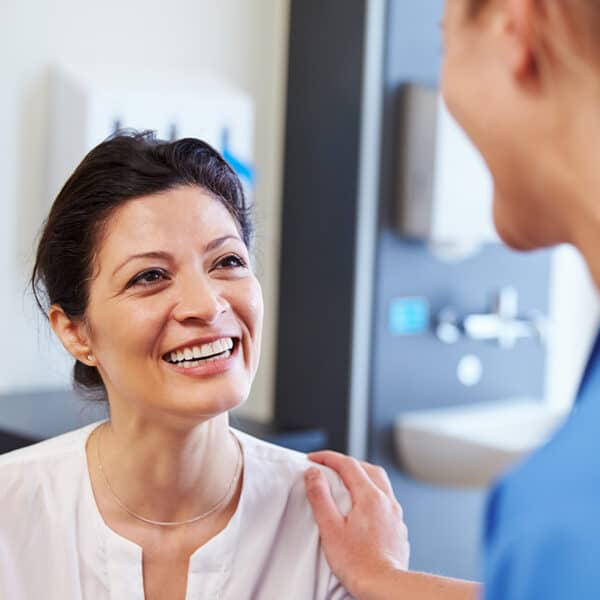 A smiling woman in a white blouse sitting in a medical setting, with a healthcare professional touching her shoulder in a supportive gesture.