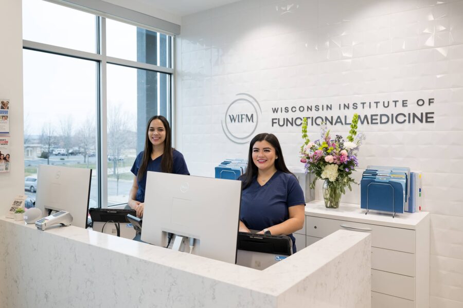 Two smiling receptionists in navy scrubs standing behind a modern white marble counter at the Wisconsin Institute of Functional Medicine (WIFM). The space features bright natural light, neatly organized files, and a vase of colorful flowers, creating a welcoming and professional environment.