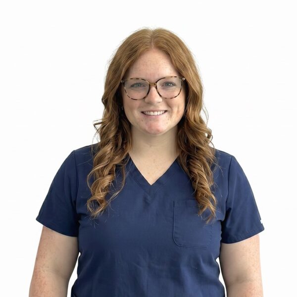 A woman with long brown hair and glasses, smiling while wearing blue scrubs against a white background.