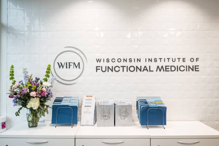 The reception counter at the Wisconsin Institute of Functional Medicine (WIFM), showcasing their logo on a sleek white textured wall. The counter features neatly organized blue file holders, informational brochures, branded gift bags, and a vibrant bouquet of fresh flowers, creating a clean and professional atmosphere.