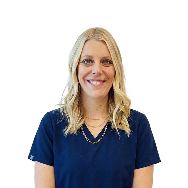 A woman with long brown hair and glasses, smiling while wearing blue scrubs against a white background.