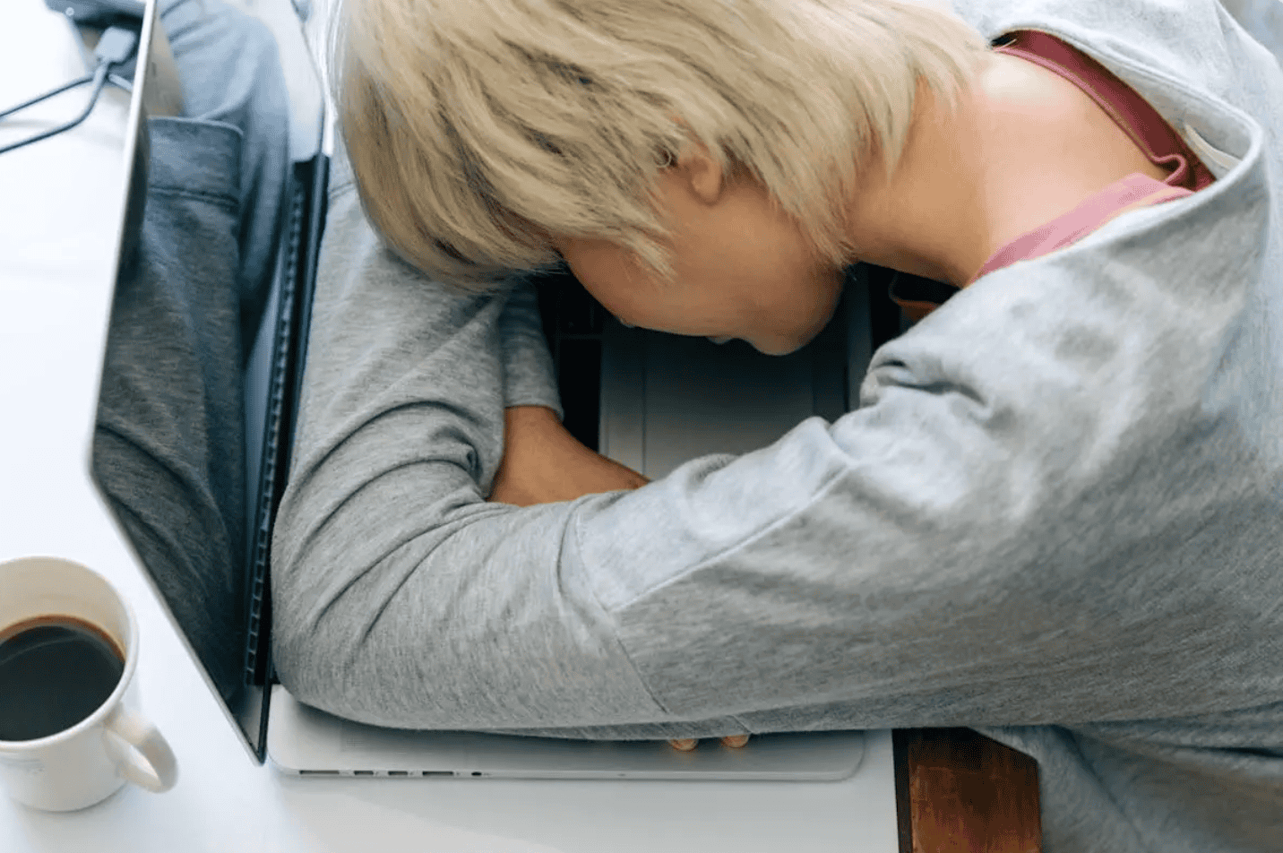 A person with short blonde hair resting their head on a laptop keyboard, with a cup of coffee nearby, appearing tired or exhausted.