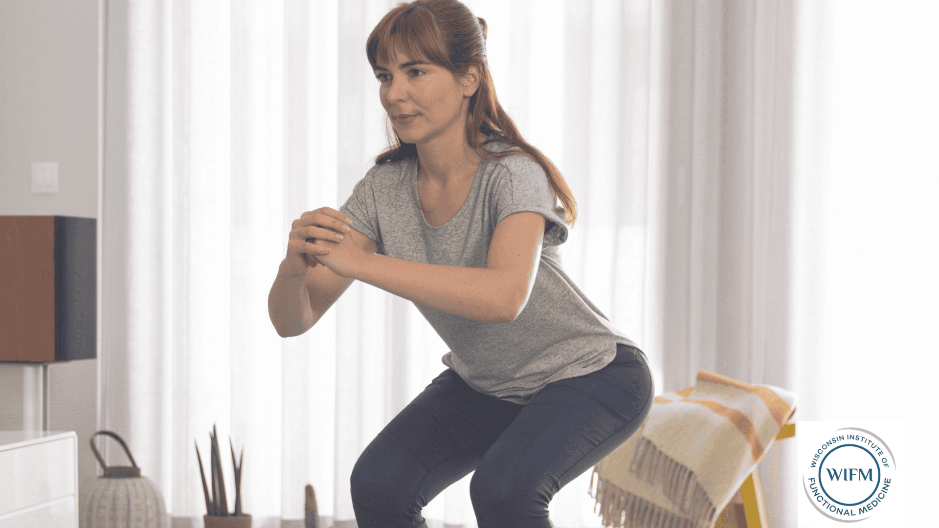 A woman in a gray shirt and dark pants performing a squat exercise in a bright living room, with the Wisconsin Institute of Functional Medicine logo in the corner.