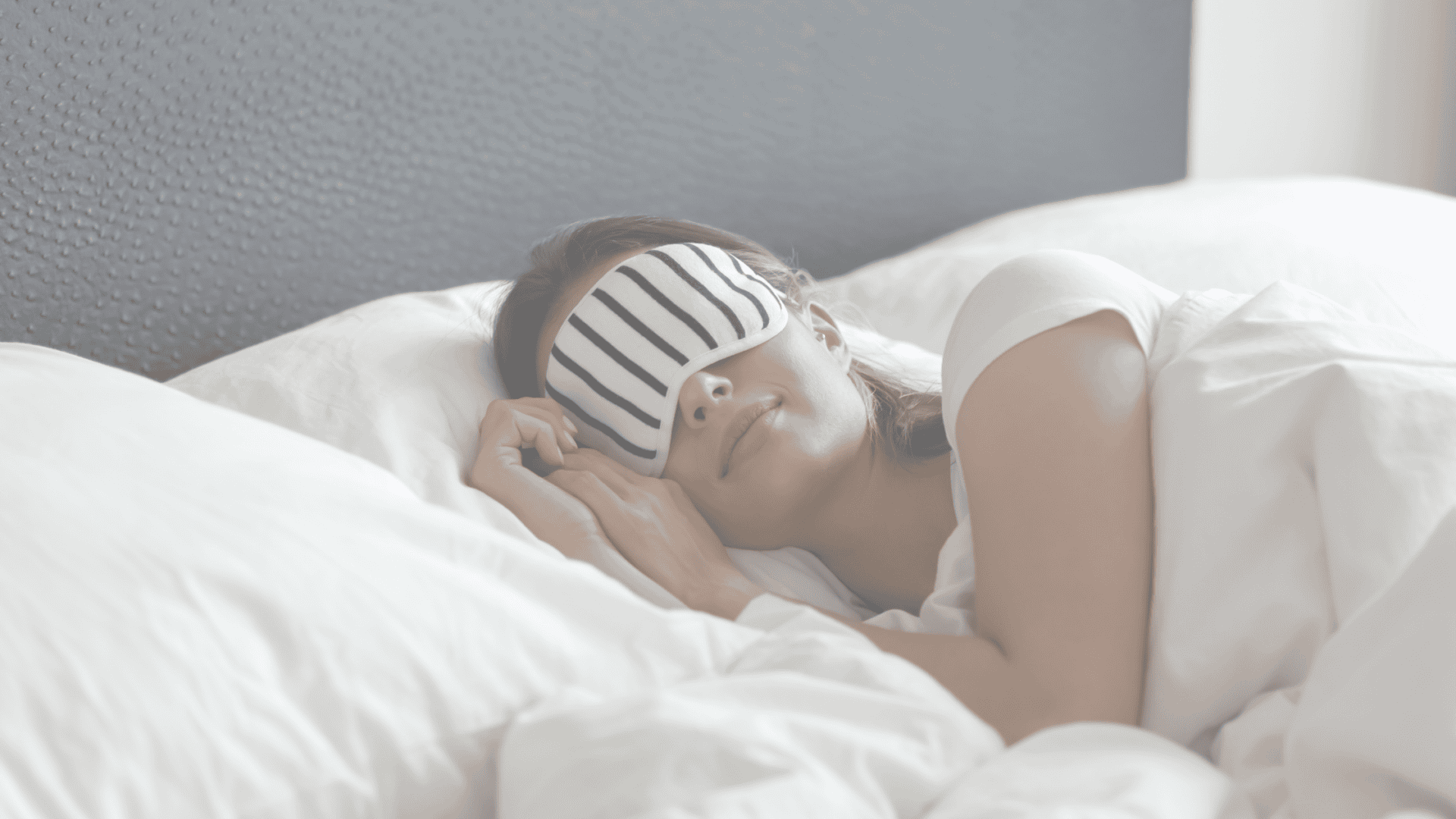 A woman lying in bed with white sheets, wearing a striped sleep mask and looking relaxed.