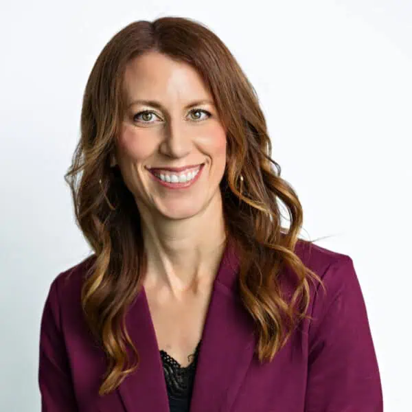 A woman with medium-length brown hair, wearing a purple top and smiling, standing in front of a white background.