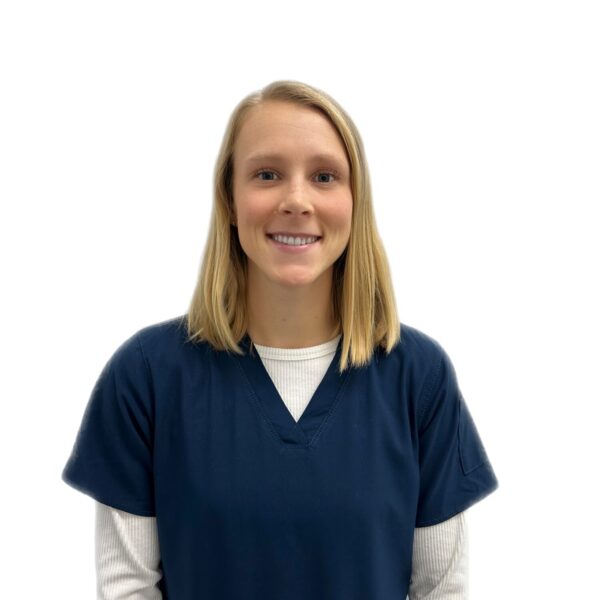 A woman with long brown hair and glasses, smiling while wearing blue scrubs against a white background.