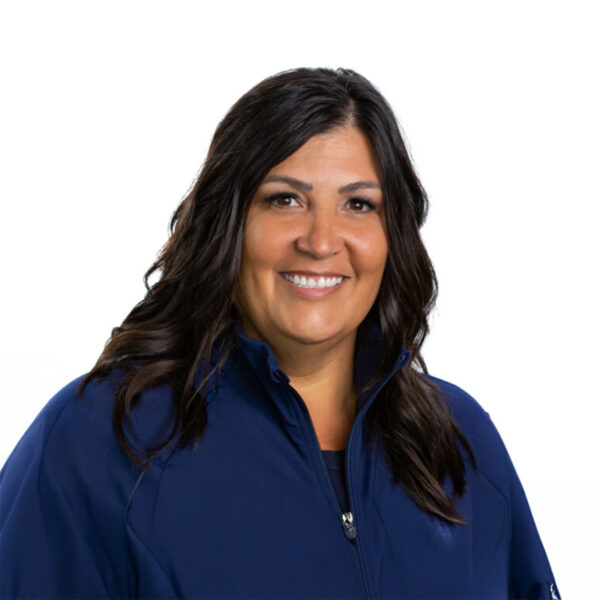 A woman with long brown hair and glasses, smiling while wearing blue scrubs against a white background.