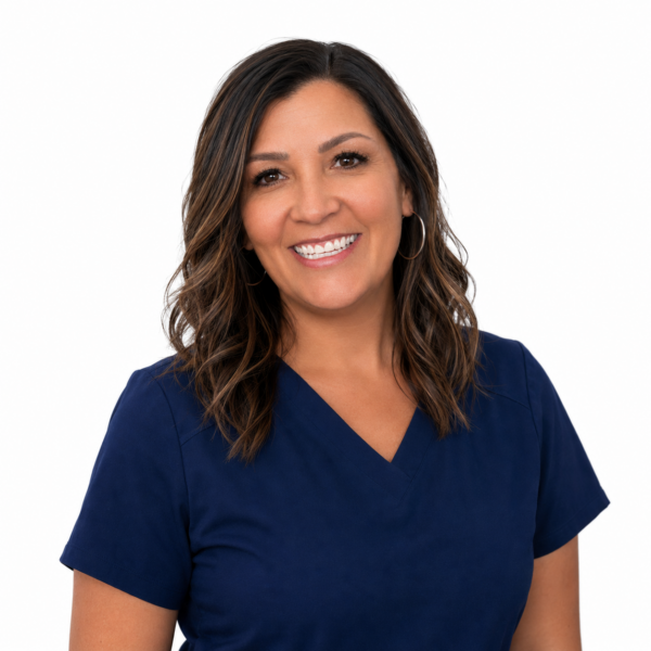 A woman with long brown hair and glasses, smiling while wearing blue scrubs against a white background.