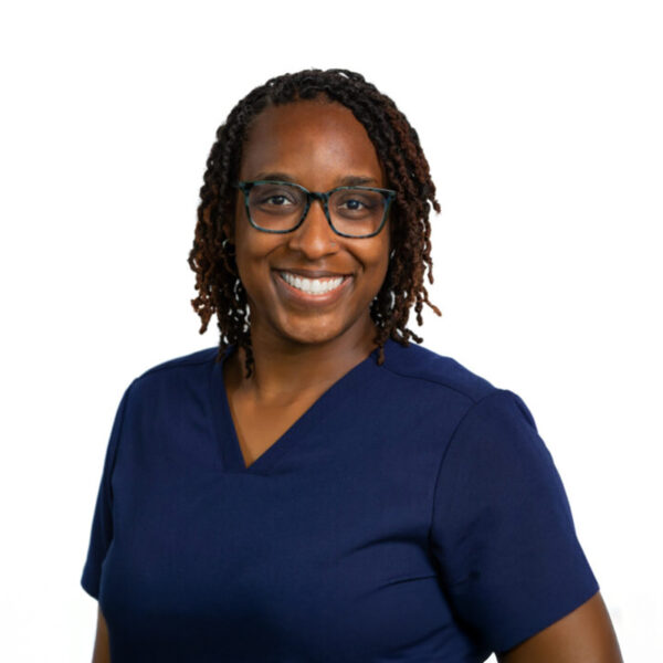 A woman with long brown hair and glasses, smiling while wearing blue scrubs against a white background.
