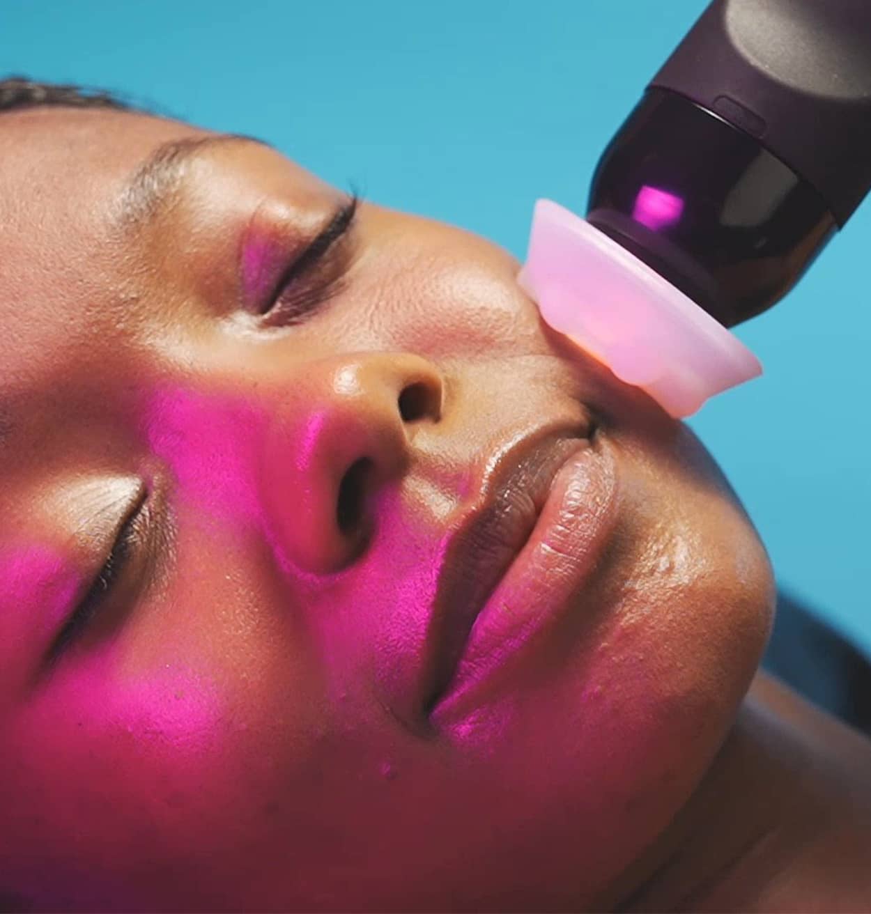 A tall woman with long brown hair holding a Glo2Facial machine, smiling confidently in a skincare clinic.