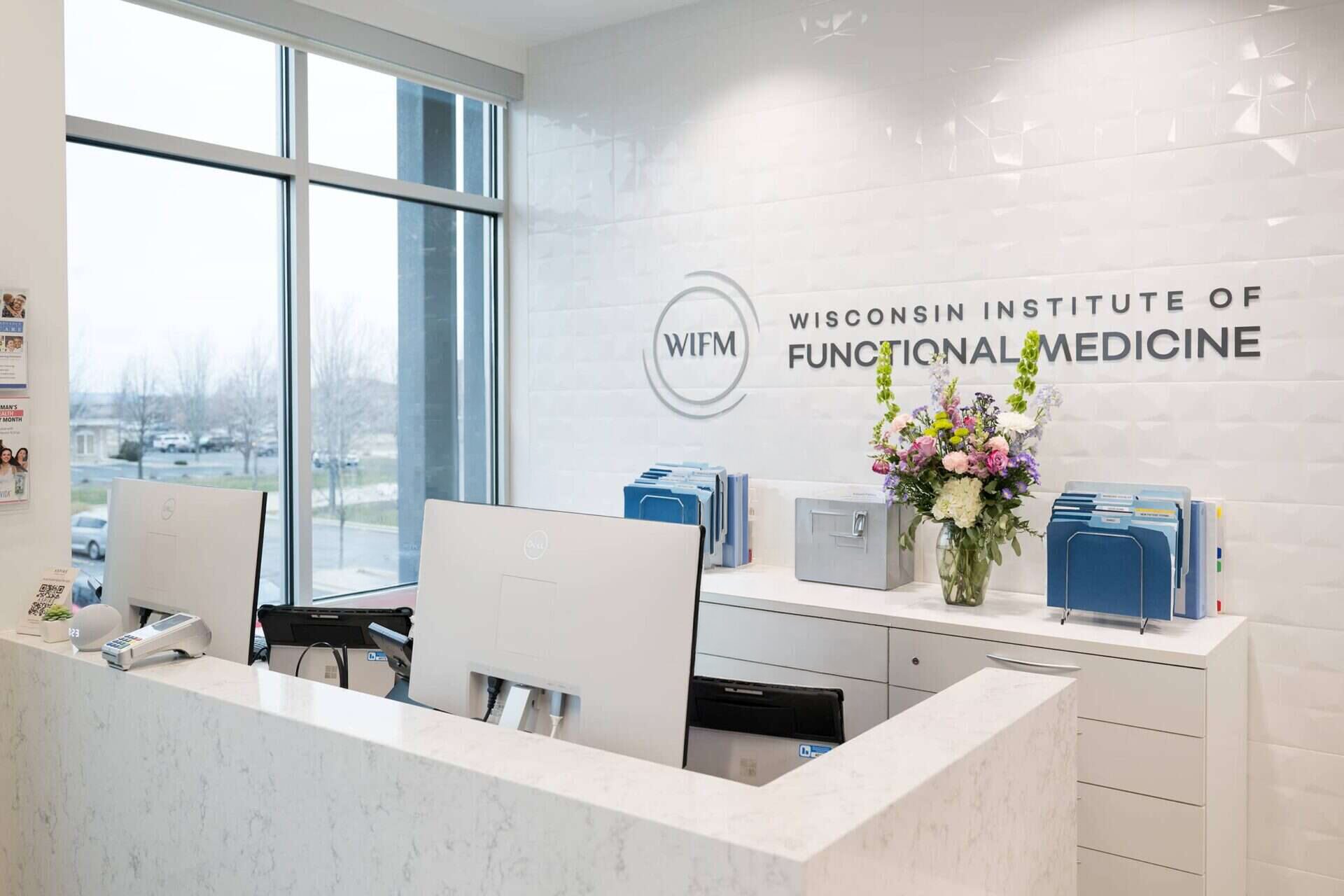 Two women with dark hair, wearing navy blue scrubs, smiling while standing at the receptionist desk in a bright white room with large windows.
