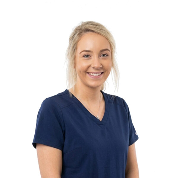 A woman with long brown hair and glasses, smiling while wearing blue scrubs against a white background.